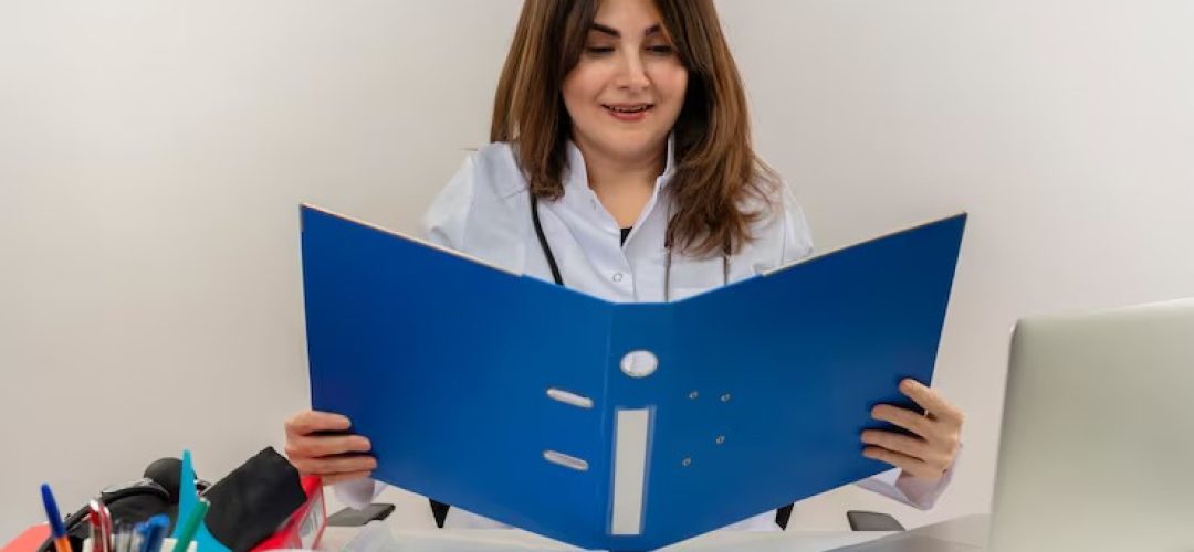 imgi_27_pleased-middle-aged-female-doctor-wearing-wearing-medical-robe-with-stethoscope-sitting-desk-work-laptop-with-medical-tools-holding-looking-folder-white-wall_141793-23496